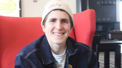 White college student sits in a red wing chair. He is wearing a beanie cap and a dark blue jacket, holding a coffee cup.