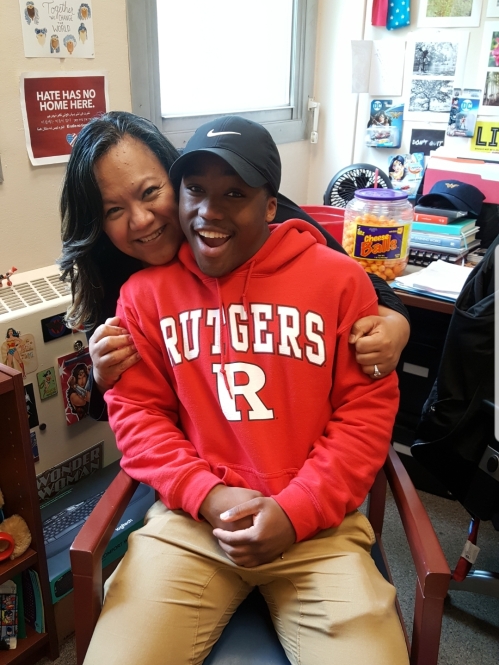 Black student wearing a red Rutgers sweatshirt and black baseball cap sits in a chair in an office. From behind a woman with black hair gives him a hug.