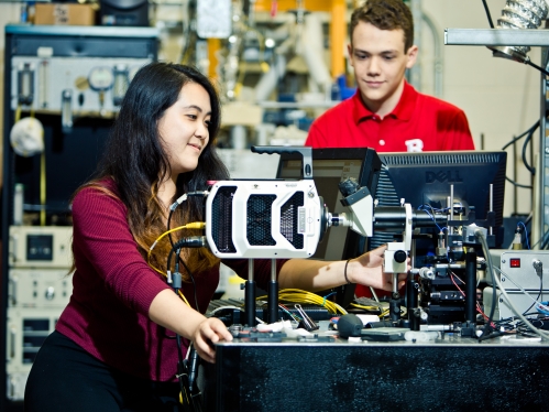 An Asian female student with long black hair, wearing maroon long sleeve shirt and a male white student wearing safety goggles and a red polo shirt conducting research in an engineering lab.